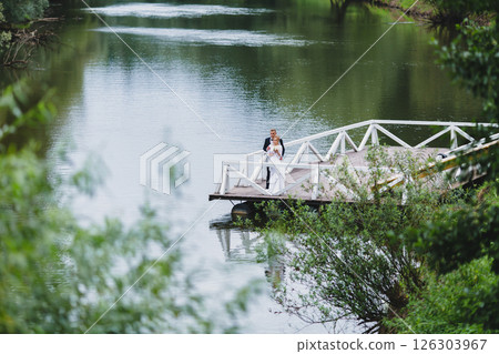 Beautiful newlywed couple in love posing on wooden pier with river in background Beautiful newlywed couple in love posing on wooden pier with river in background 126303967