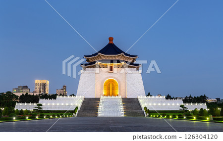 Evening view of the grand Chiang Kai-shek Memorial Hall in Taipei, Taiwan, illuminated against a clear blue sky. The imposing white marble structure with its blue octagonal roof stands atop a wide 126304120