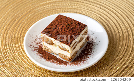 A slice of classic Tiramisu dessert on a white plate, dusted with cocoa, sits on a golden textured placemat. 126304209