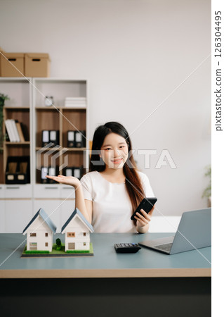 Young real estate agent worker working with laptop and tablet at table in office and small house beside it. Young real estate agent worker working with laptop and tablet at table in office and small house beside it. 126304495