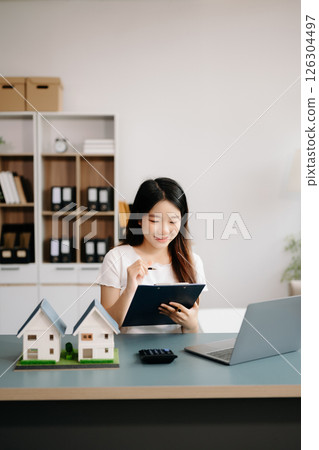 Young real estate agent worker working with laptop and tablet at table in office and small house beside it. Young real estate agent worker working with laptop and tablet at table in office and small house beside it. 126304497