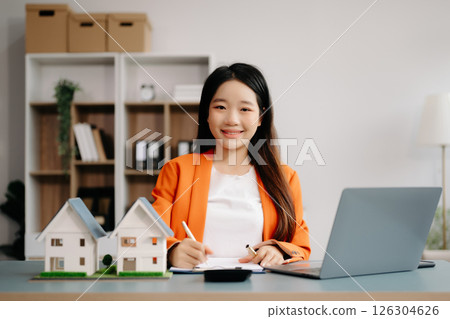 Young real estate agent worker working with laptop and tablet at table in office and small house beside it. Young real estate agent worker working with laptop and tablet at table in office and small house beside it. 126304626