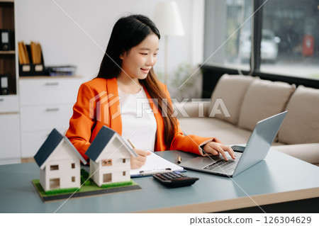 Young real estate agent worker working with laptop and tablet at table in office and small house beside it. Young real estate agent worker working with laptop and tablet at table in office and small house beside it. 126304629