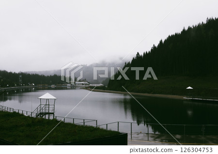 A scenic view of a calm lake nestled within majestic mountains on a cloudy foggy day. A serene pond surrounded by lush mountains A tranquil atmosphere A scenic view of a calm lake nestled within majestic mountains on a cloudy foggy day. A serene pond surrounded by lush mountains A tranquil atmosphere 126304753