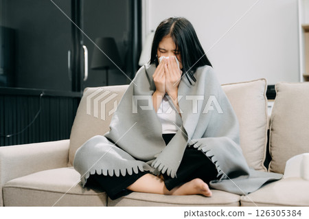 Young Asian woman suffering from flu symptoms, covered with a blanket on a sofa. Concept of illness, healthcare, Young Asian woman suffering from flu symptoms, covered with a blanket on a sofa. Concept of illness, healthcare, 126305384