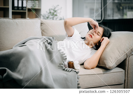 Young Asian woman suffering from flu symptoms, covered with a blanket on a sofa. Concept of illness, healthcare Young Asian woman suffering from flu symptoms, covered with a blanket on a sofa. Concept of illness, healthcare 126305507