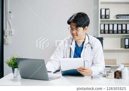 Confident young male doctor in white medical uniform sit at desk working on computer. 126305825