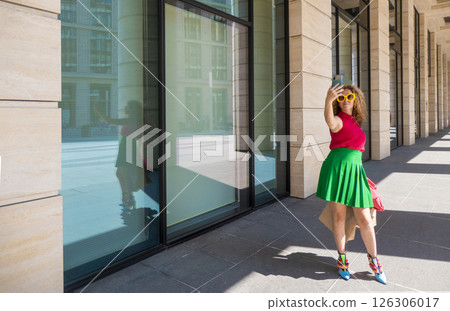 a beautiful young woman in bright clothes with a phone and bags takes selfies or takes pictures of the city on a walk a beautiful young woman in bright clothes with a phone and bags takes selfies or takes pictures of the city on a walk 126306017