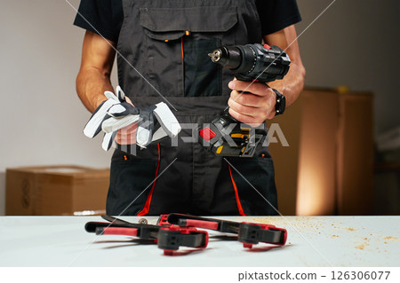 Person in work overalls holding cordless power drill and safety gloves at workbench with tools and wood shavings 126306077