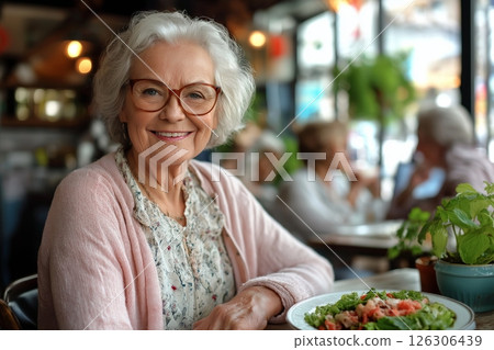 White-haired woman wearing a floral blouse and pink cardigan smiling near a plate of greens 126306439