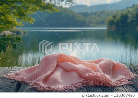 A pink blanket with fringes lies gently on a rustic wooden dock beside a calm forest lake 126307480