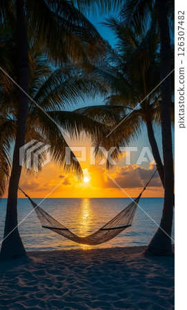 A hammock stretches between palm trees on the beach, glowing under a fiery tropical sunset sky 126307482