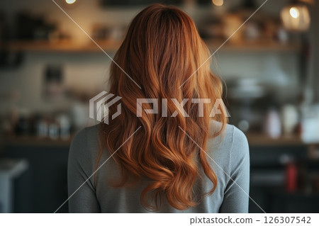 A woman with voluminous red waves viewed from behind in a cozy room with warm tones and bokeh 126307542