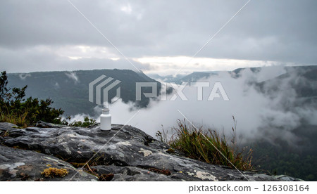 A white bottle sits on a rock overlooking a misty mountain valley under a cloudy sky, creating a serene and atmospheric scene. A white bottle sits on a rock overlooking a misty mountain valley under a cloudy sky, creating a serene and atmospheric scene. 126308164
