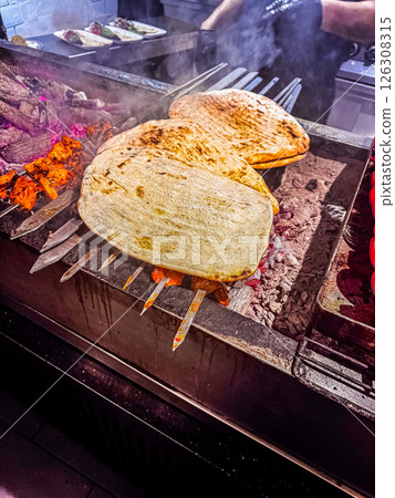 Flatbreads are being grilled on skewers over hot coals in a lively kitchen setting. High quality photo 126308315