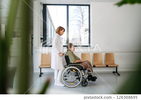 Friendly doctor pushing elderly patient in wheelchair across hospital hallway, 126308695