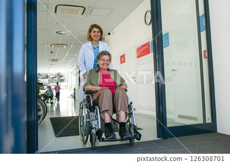 Friendly doctor pushing elderly patient in wheelchair across hospital hallway, Friendly doctor pushing elderly patient in wheelchair across hospital hallway, 126308701