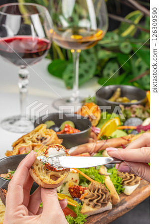 Spreading soft cheese on toasted bread with gourmet appetizers and wine in the background 126309590