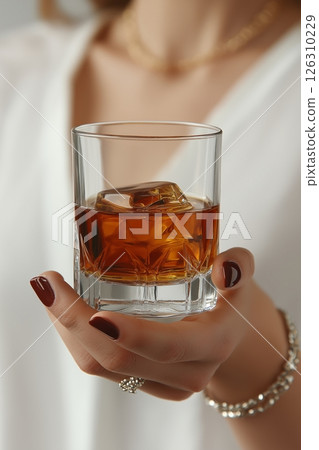 A womans hand holding a whiskey glass with amber liquid and ice cubes, adorned with jewelry A womans hand holding a whiskey glass with amber liquid and ice cubes, adorned with jewelry 126310229