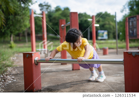 Asian Young girl exercising outdoors on a playground using a fitness bar to strengthen her body and promote health in a vibrant green environment Asian Young girl exercising outdoors on a playground using a fitness bar to strengthen her body and promote health in a vibrant green environment 126311534
