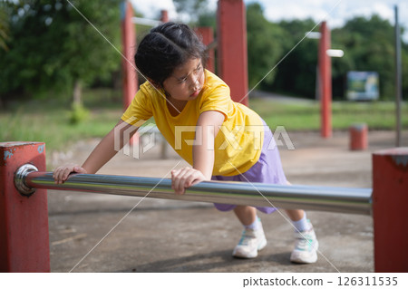 Asian Young girl practicing strength training on playground equipment in a sunny park, focusing on fitness and outdoor activities with determination and joy. 126311535