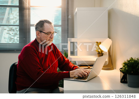 Senior man in red sweater working online in a calm room with natural and lamp light 126311848