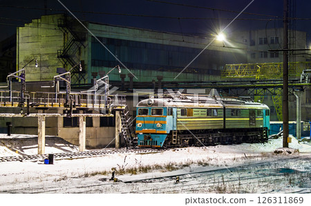 Nighttime view of an old Soviet locomotive parked on snowy tracks at the Lviv Locomotive Repairing Plant in Ukraine. The cold industrial scene features concrete structures, a weathered locomotive with 126311869