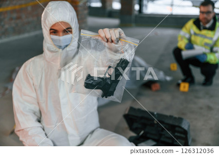 Woman is holding pistol that is in the bag. Detectives are collecting evidence in a crime scene near dead body 126312036