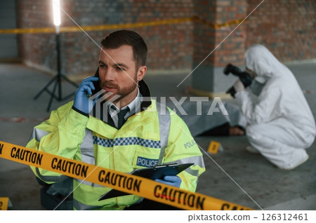 Man is talking by phone. Detectives are collecting evidence in a crime scene near dead body Man is talking by phone. Detectives are collecting evidence in a crime scene near dead body 126312461