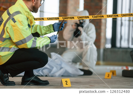 Woman is using camera, making photos. Detectives are collecting evidence in a crime scene near dead body 126312480