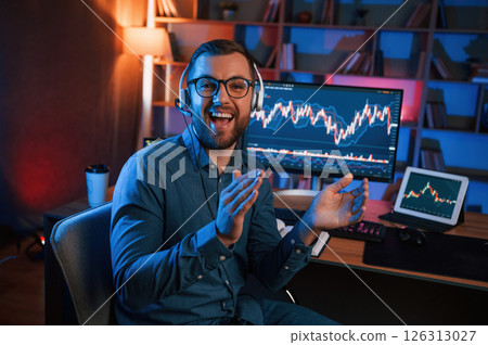 Sitting and looking at the camera. Stock trader is working by pc indoors. Conception of exchange and money 126313027