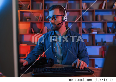 In glasses. Man is sitting by a computer indoors at home. Neon lighting 126313448