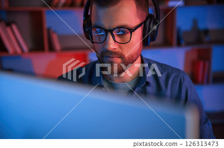 Front view. Man is sitting by a computer indoors at home. Neon lighting 126313473