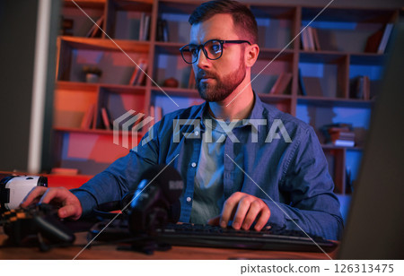 Front view. Man is sitting by a computer indoors at home. Neon lighting Front view. Man is sitting by a computer indoors at home. Neon lighting 126313475