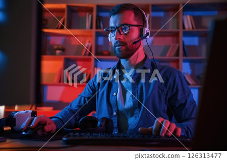 Front view. Man is sitting by a computer indoors at home. Neon lighting Front view. Man is sitting by a computer indoors at home. Neon lighting 126313477