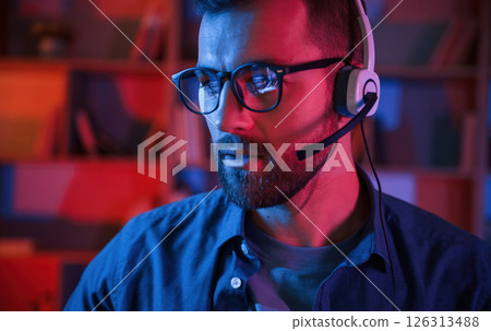 Portrait of man that is sitting by a computer indoors at home. Neon lighting Portrait of man that is sitting by a computer indoors at home. Neon lighting 126313488