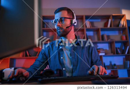 Concentrated man is sitting by a computer indoors at home. Neon lighting Concentrated man is sitting by a computer indoors at home. Neon lighting 126313494