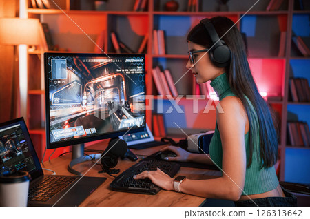Side view. Young pretty female gamer is playing shooter. Neon lighting Side view. Young pretty female gamer is playing shooter. Neon lighting 126313642