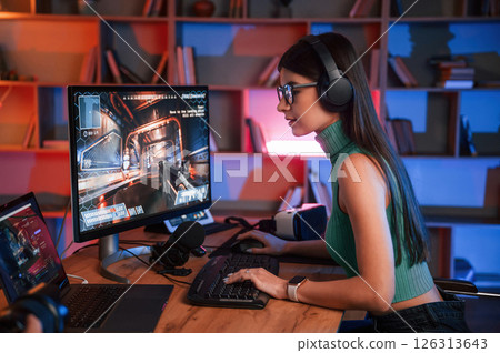 Side view. Young pretty female gamer is playing shooter. Neon lighting Side view. Young pretty female gamer is playing shooter. Neon lighting 126313643