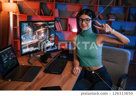 Smiling, showing victory gesture. Young pretty female gamer is playing shooter. Neon lighting 126313650