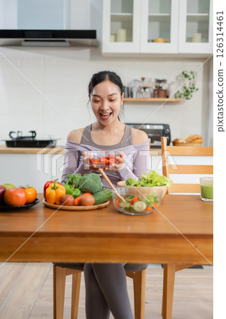 Fit woman enjoys healthy fresh vegetables and fruits breakfast sitting in the kitchen. 126314461