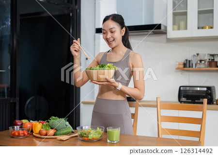 Fit woman enjoys healthy fresh vegetables and fruits breakfast sitting in the kitchen. 126314492