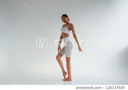 In white fitness clothes. Young caucasian woman with slim body shape is indoors in the studio 126314585