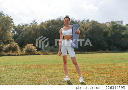 Standing with yoga mat. Young caucasian woman with slim body shape is in the fitness clothes outdoors Standing with yoga mat. Young caucasian woman with slim body shape is in the fitness clothes outdoors 126314595