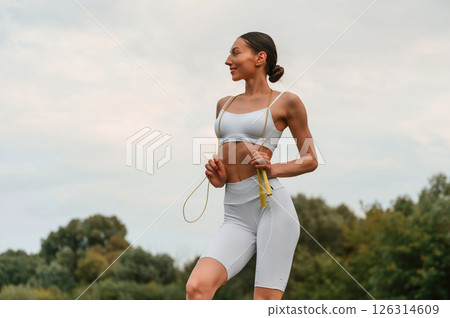 Holding jumping rope. Young caucasian woman with slim body shape is in the fitness clothes outdoors Holding jumping rope. Young caucasian woman with slim body shape is in the fitness clothes outdoors 126314609