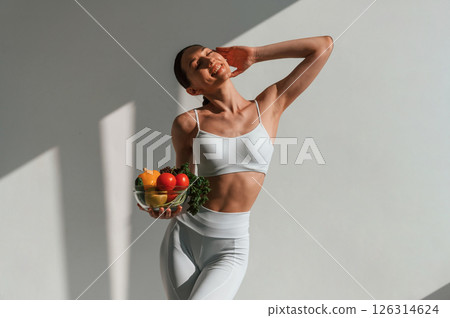 Holding a bowl of vegetables and fruits. Young caucasian woman with slim body shape is indoors in the studio Holding a bowl of vegetables and fruits. Young caucasian woman with slim body shape is indoors in the studio 126314624