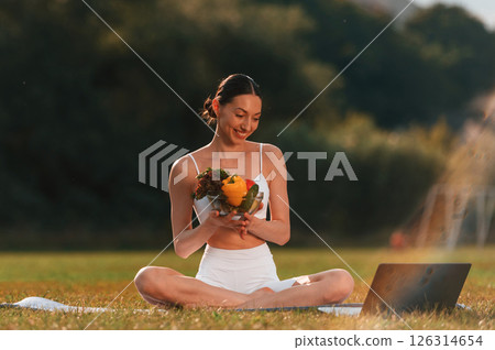 Watching at laptop, holding vegetables. Young caucasian woman with slim body shape is in the fitness clothes outdoors 126314654