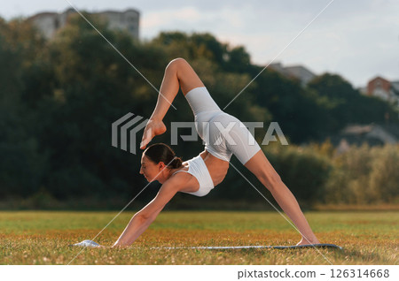 Side view. Doing yoga. Young caucasian woman with slim body shape is in the fitness clothes outdoors Side view. Doing yoga. Young caucasian woman with slim body shape is in the fitness clothes outdoors 126314668
