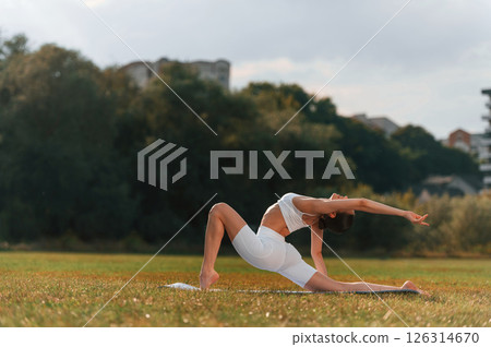 Side view. Doing yoga. Young caucasian woman with slim body shape is in the fitness clothes outdoors Side view. Doing yoga. Young caucasian woman with slim body shape is in the fitness clothes outdoors 126314670
