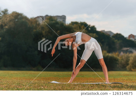 Standing on the mat, doing stretching. Young caucasian woman with slim body shape is in the fitness clothes outdoors 126314675